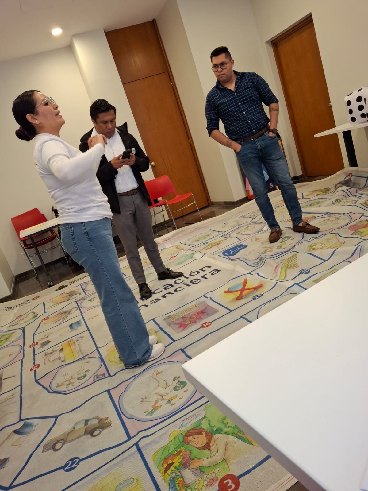 Tres personas jugando un juego de mesa gigante en el suelo con paneles ilustrados y caminos, dentro de un espacio cerrado.