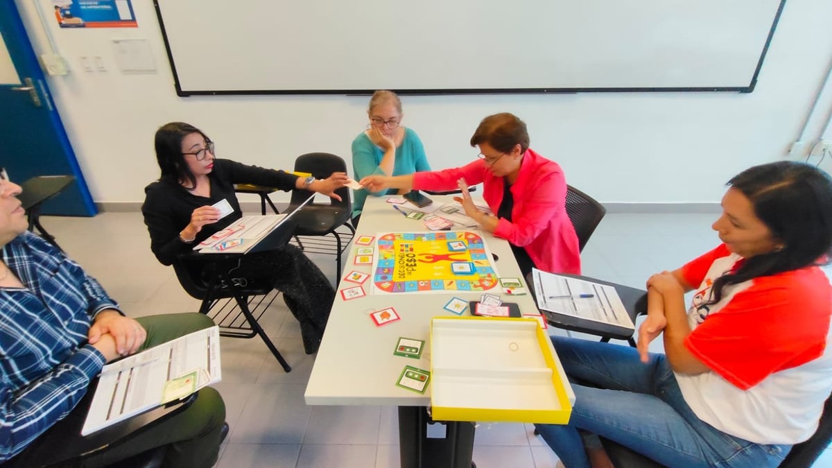 Grupo de estudiantes y una instructora alrededor de una mesa con un juego de mesa durante una actividad en aula.