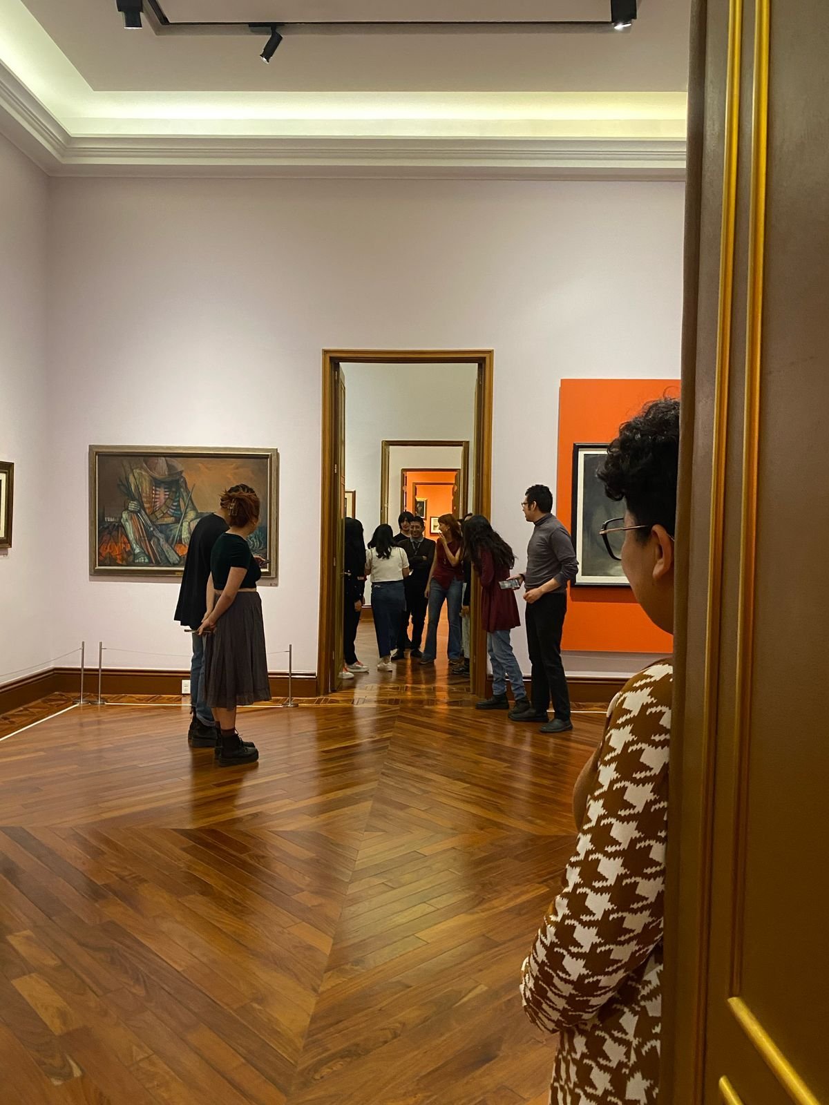 Visitantes observando obras en una sala de galería con muros blancos y piso de madera