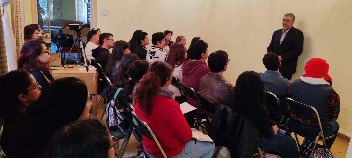 Grupo de personas sentadas en círculo durante una presentación, con un expositor de pie dirigiéndose al público.