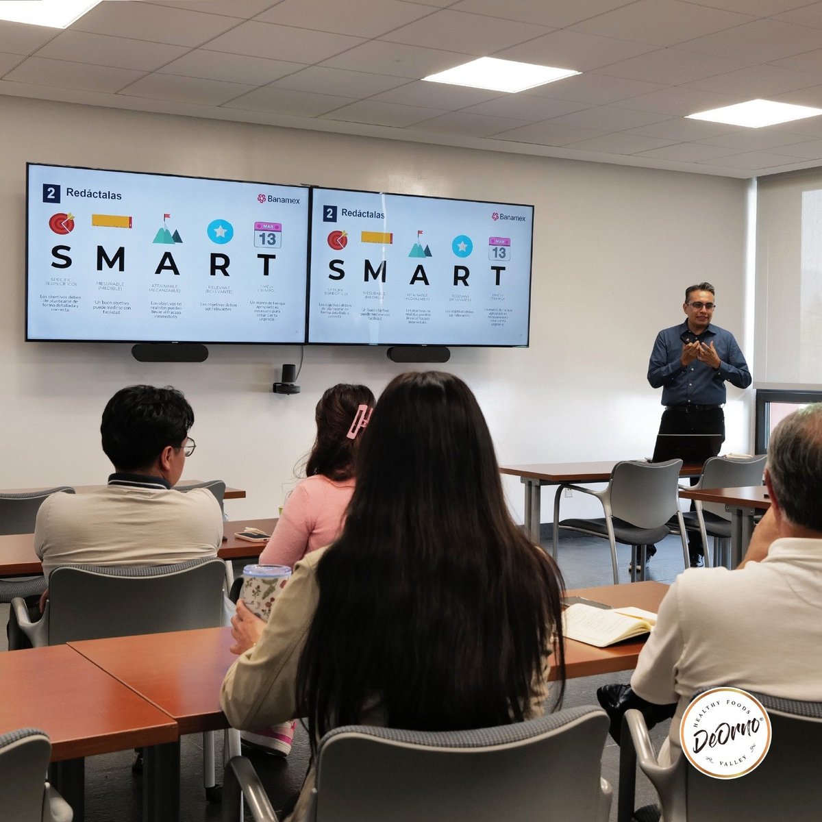 Capacitación en sala de juntas con un facilitador presentando objetivos SMART frente a dos pantallas Banamex, ante un grupo pequeño de asistentes.