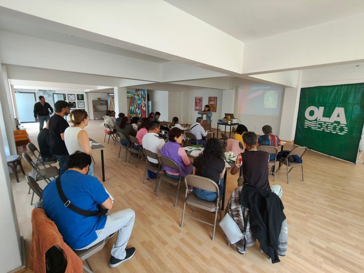 Sala con asistentes sentados frente a un proyector y un estand verde con la palabra OLA, durante una charla para emprendedores.