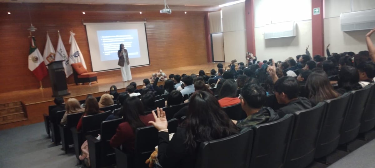 Conferencista en un auditorio con paredes de madera, ante un público numeroso con la mano levantada.